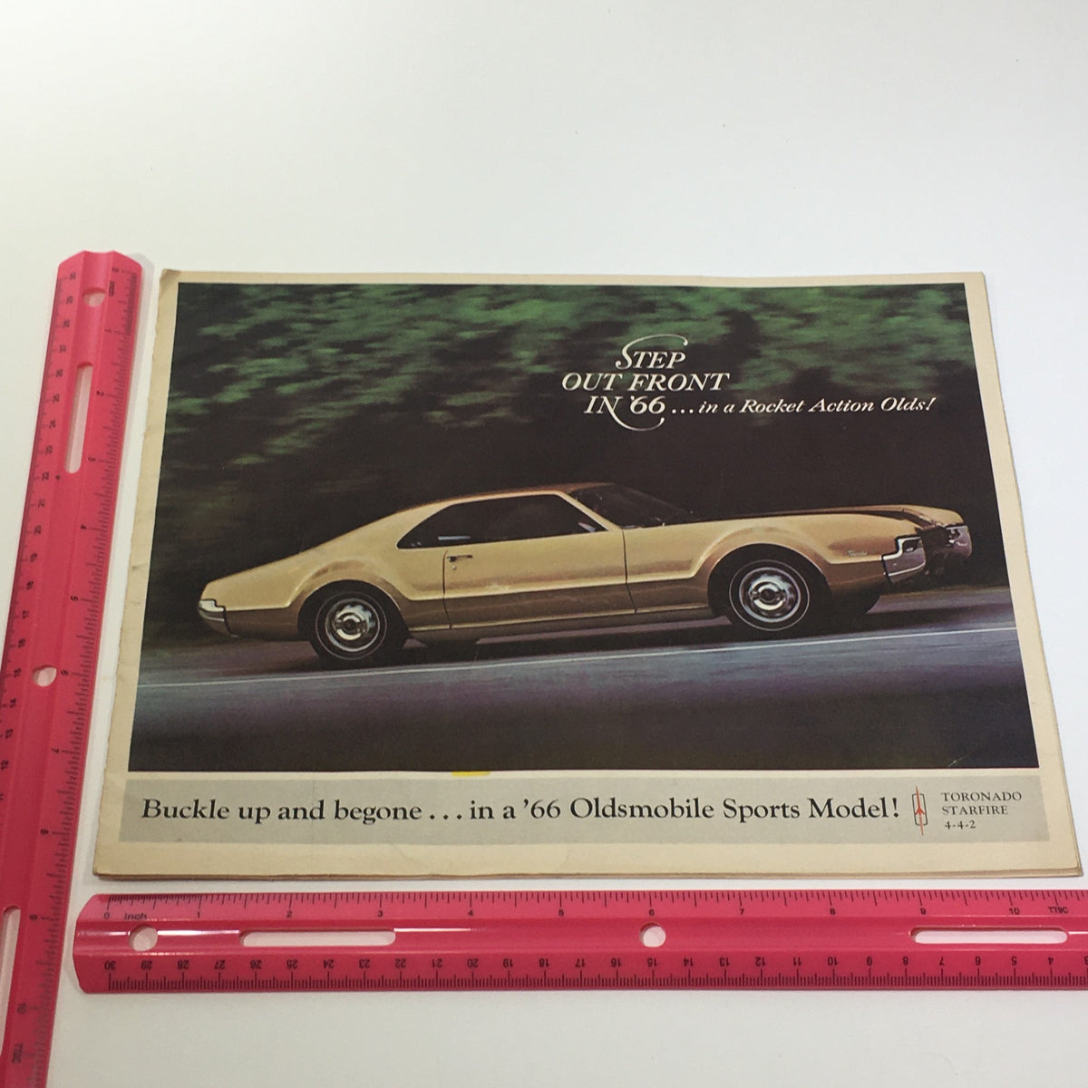 Another perspective of the 1966 Oldsmobile Sports Model Tornado-Starfire brochure cover, highlighting the gold car with a ruler for size comparison, captioned 'Buckle up and begone in a '66 Oldsmobile Sports Model!'.