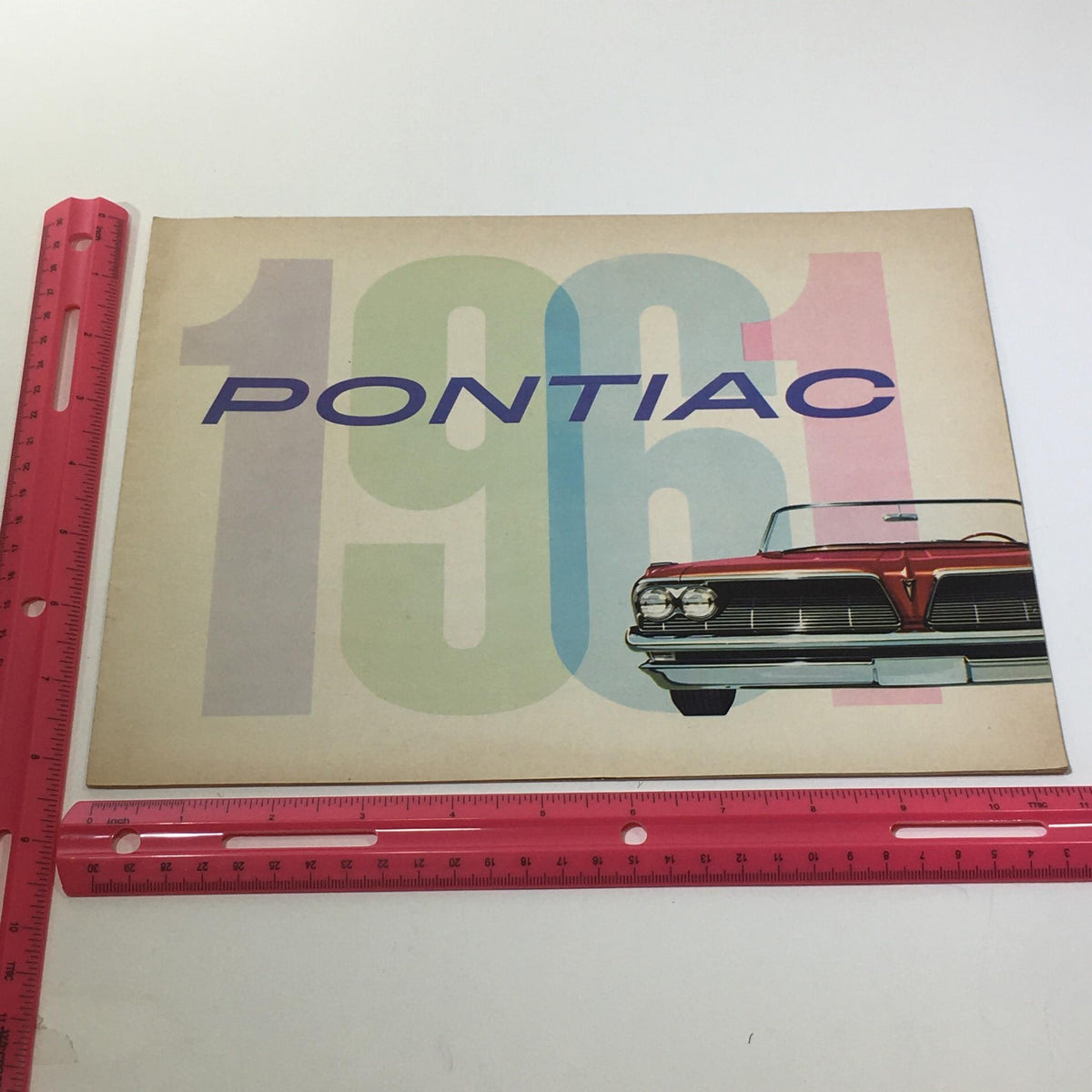 Side view of the 1961 Pontiac Catalina Sport 2-Door Coupe Type V-8 Hydra-Matic Engine Car Catalog with a ruler for scale, showcasing its colorful cover design.