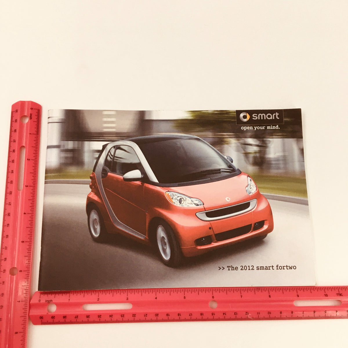Cover image of the 2012 Smart Fortwo City Solution Hill-Start Assist Car Brochure showing a red Smart Fortwo car in motion, highlighting its compact design.