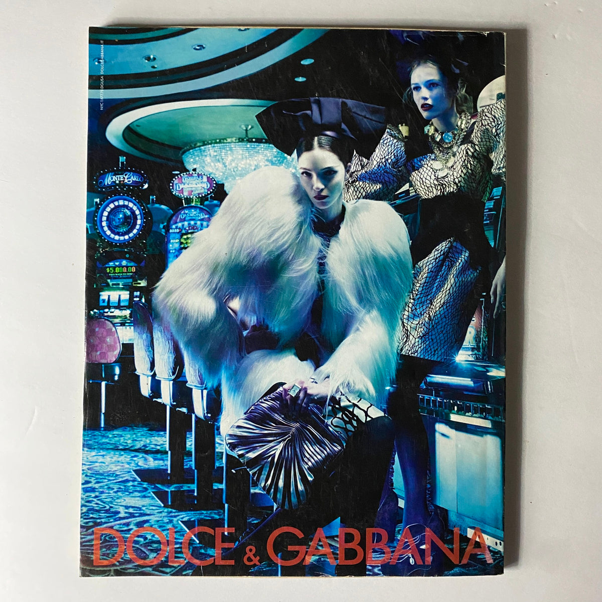 Dolce & Gabbana fashion magazine cover featuring a glamorous duo in bold outfits against a casino backdrop, highlighting vibrant fashion and luxury.