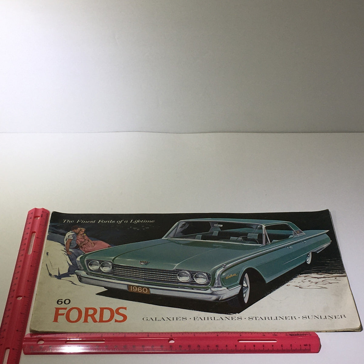 Side view of the 1960 Ford Galaxies-Fairlanes-Starliner-Sunliner V-8 Engine Car Catalog Brochure with a ruler for scale, emphasizing the impressive vintage design.