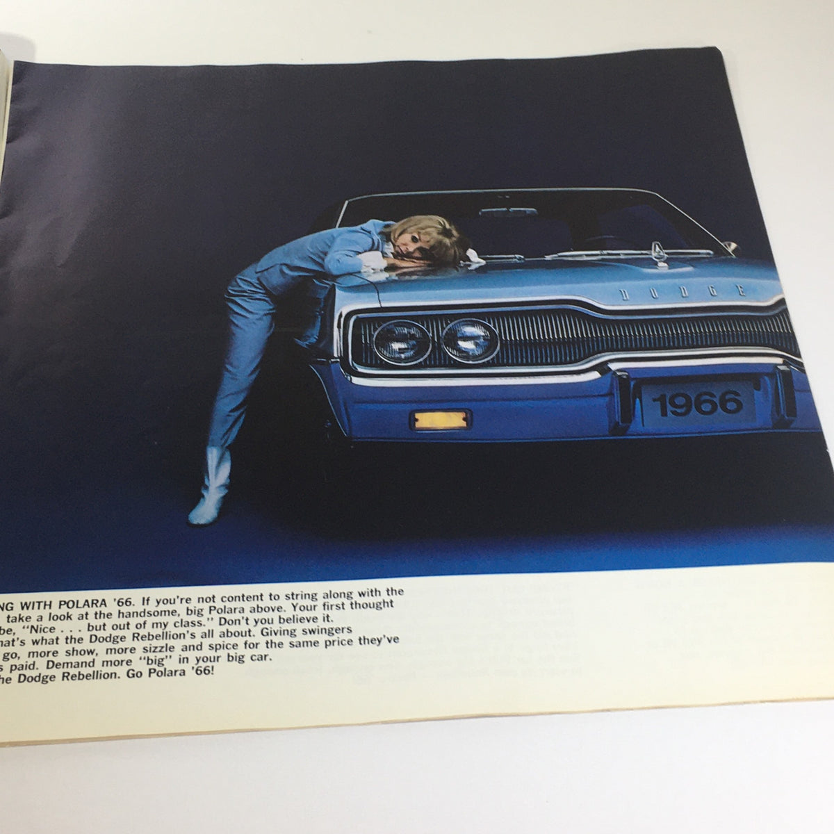 1966 Dodge Polara Monaco by Chrysler 2-Door Convertible Car Catalog Brochure showcasing a model leaning on the car's hood, against a vibrant blue backdrop.