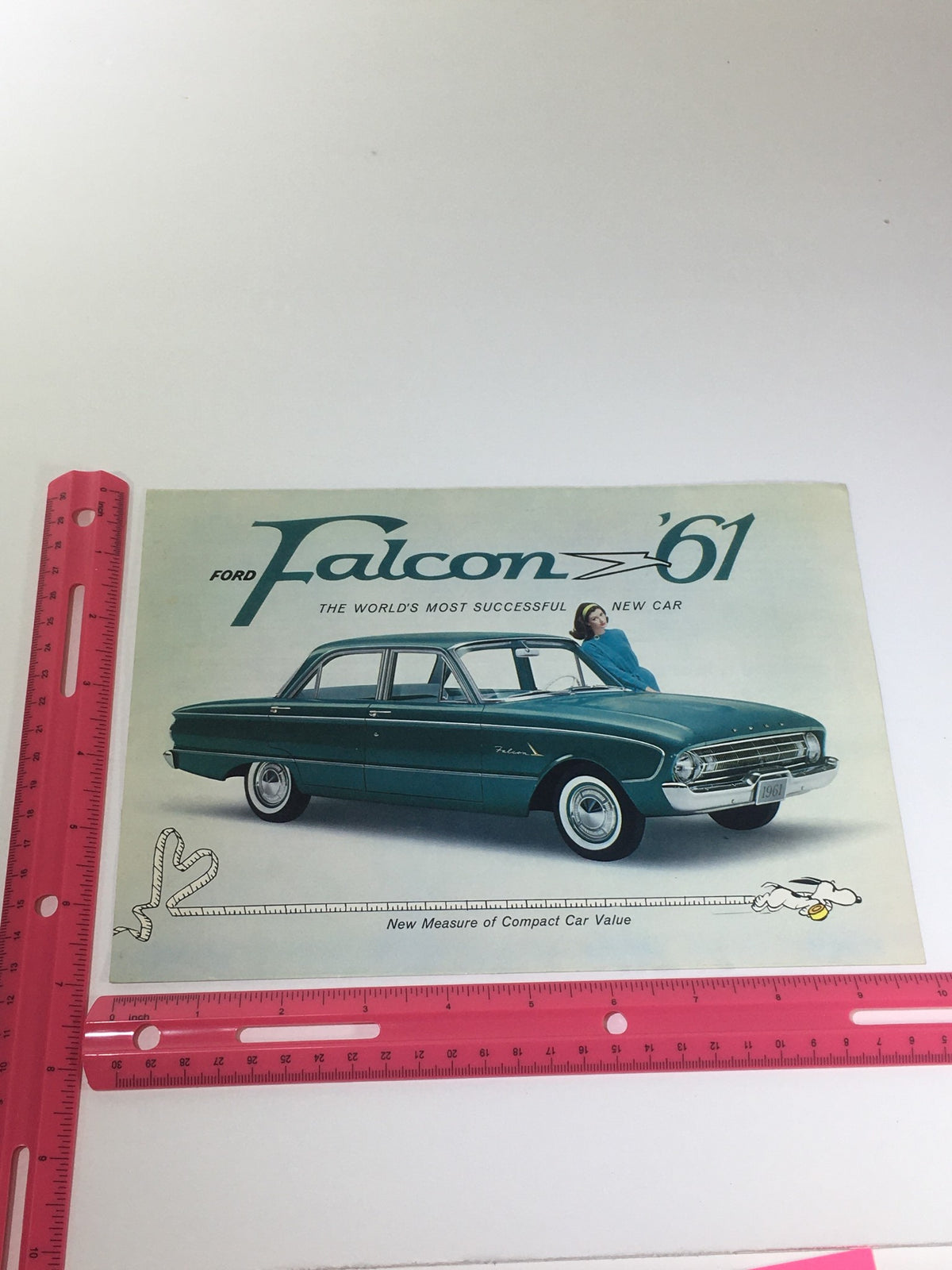 1961 Ford Falcon Fordomatic 2-Speed car catalog brochure front cover alongside a ruler for size reference, highlighting the classic design in a playful manner.
