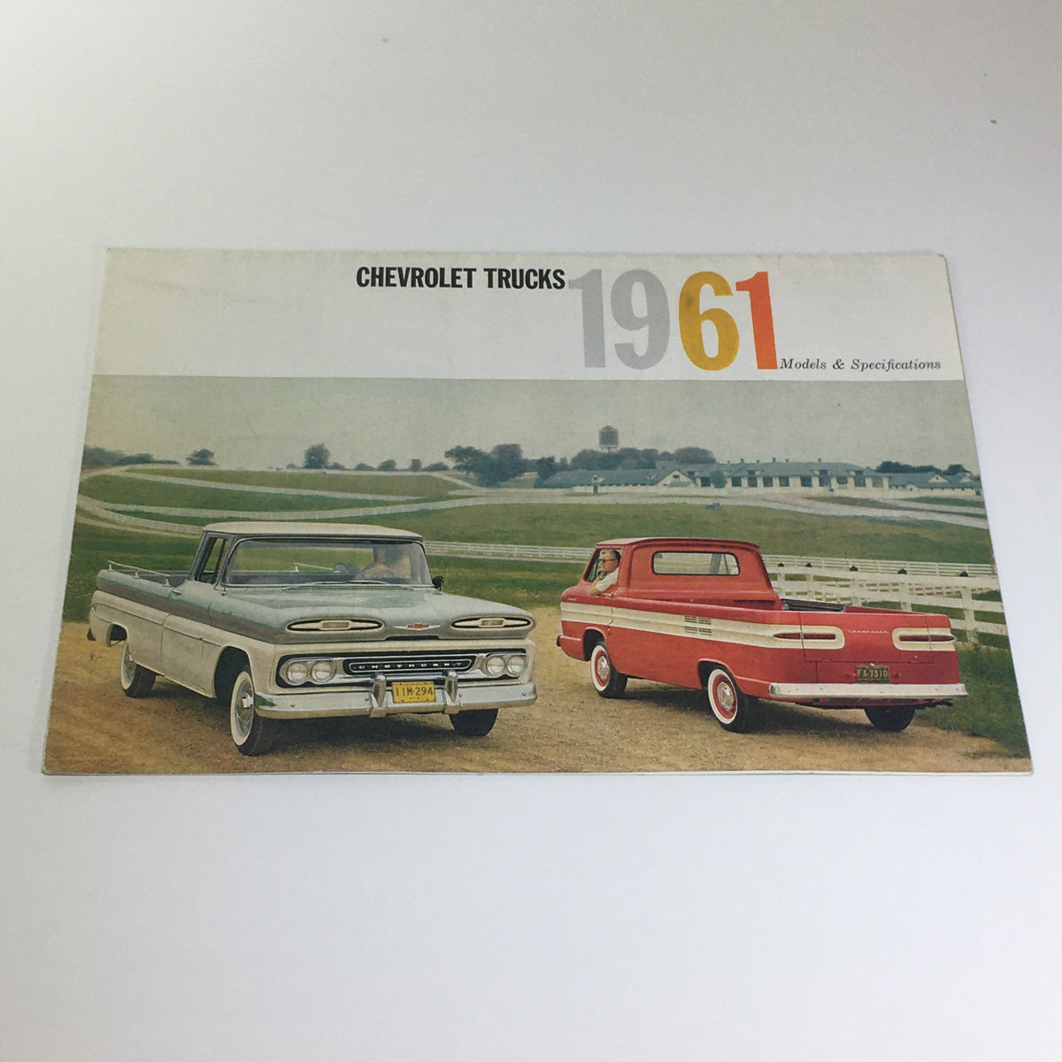 Cover of Chevrolet Trucks of 1961 Models & Specifications Light-Heavy Duty Trucks Catalog featuring a gray pickup truck and a red pickup truck parked on a rural road.