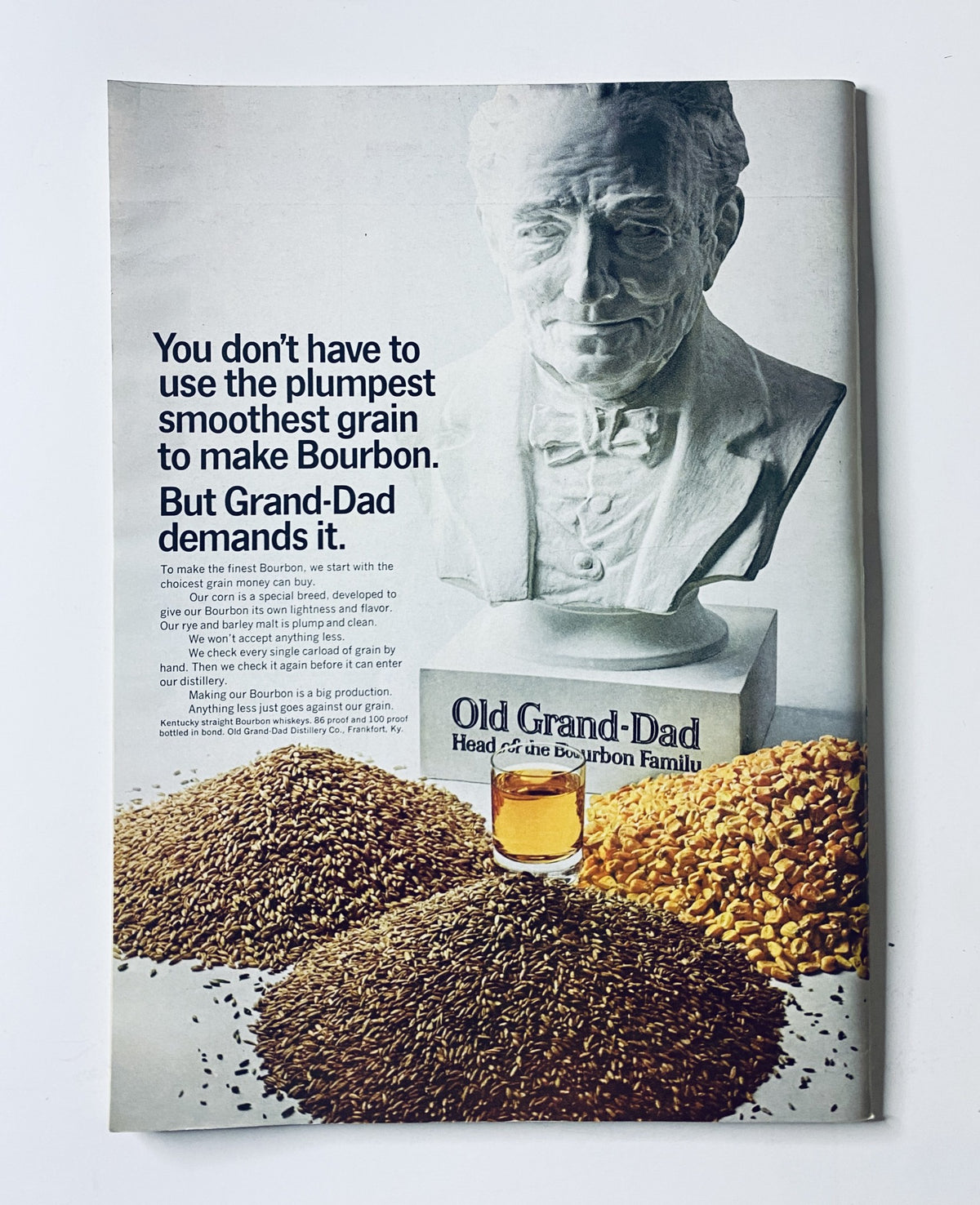 Old Grand-Dad Bourbon advertisement featuring a bust sculpture and grains, highlighting the quality ingredients for bourbon production. The ad states 'You don't have to use the plumpest smoothest grain to make Bourbon. But Grand-Dad demands it.', promoting the Old Grand-Dad brand.