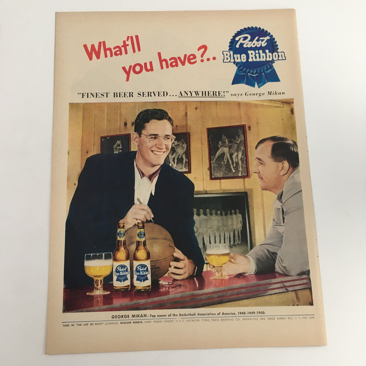 1950 Pabst Blue Ribbon Finest Beer Starring George Mikan Vintage Print Ad featuring George Mikan smiling with two bottles of Pabst Blue Ribbon beer and a basketball, in a bar setting.