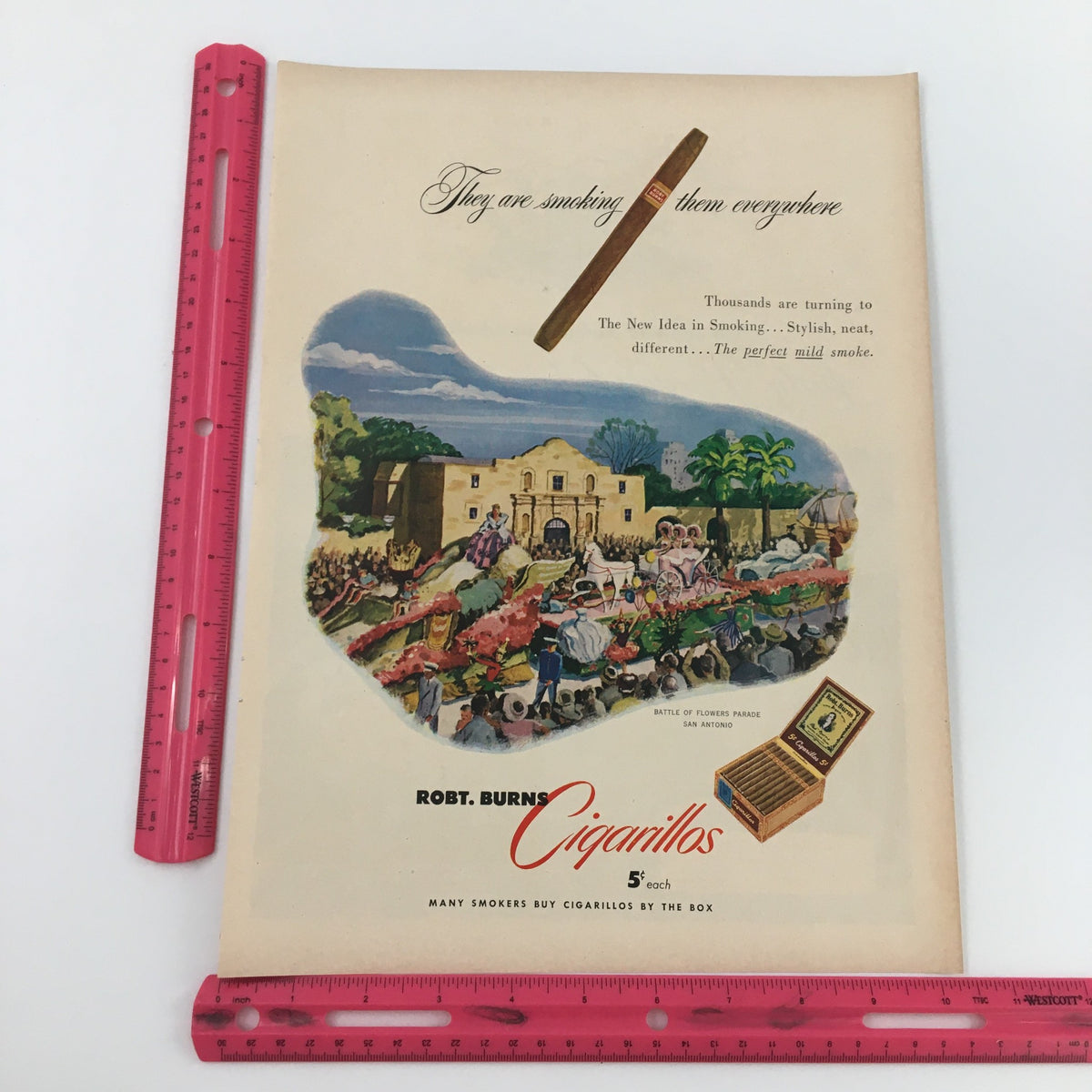 1950 Robt Burns Cigarillos By The Box Print Ad featuring vibrant artwork and a vintage design. The ad showcases a picturesque scene with a tobacco shop backdrop, highlighting the cigarillos product alongside a playful slogan about smoking.