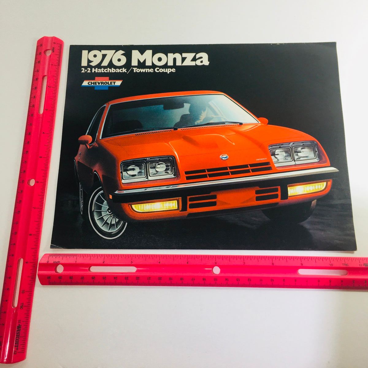 A close-up view of the Vintage 1976 Monza 2+2 Hatchback / Towne Coupe Chevrolet 2-Door Sedan Brochure with a ruler for size reference.