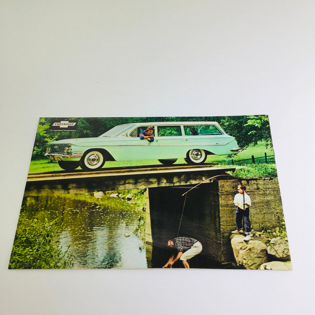 1961 Chevrolet Biscayne Series Station Wagon promotional image showing a light blue station wagon on a bridge, with a scenic background and two people fishing.