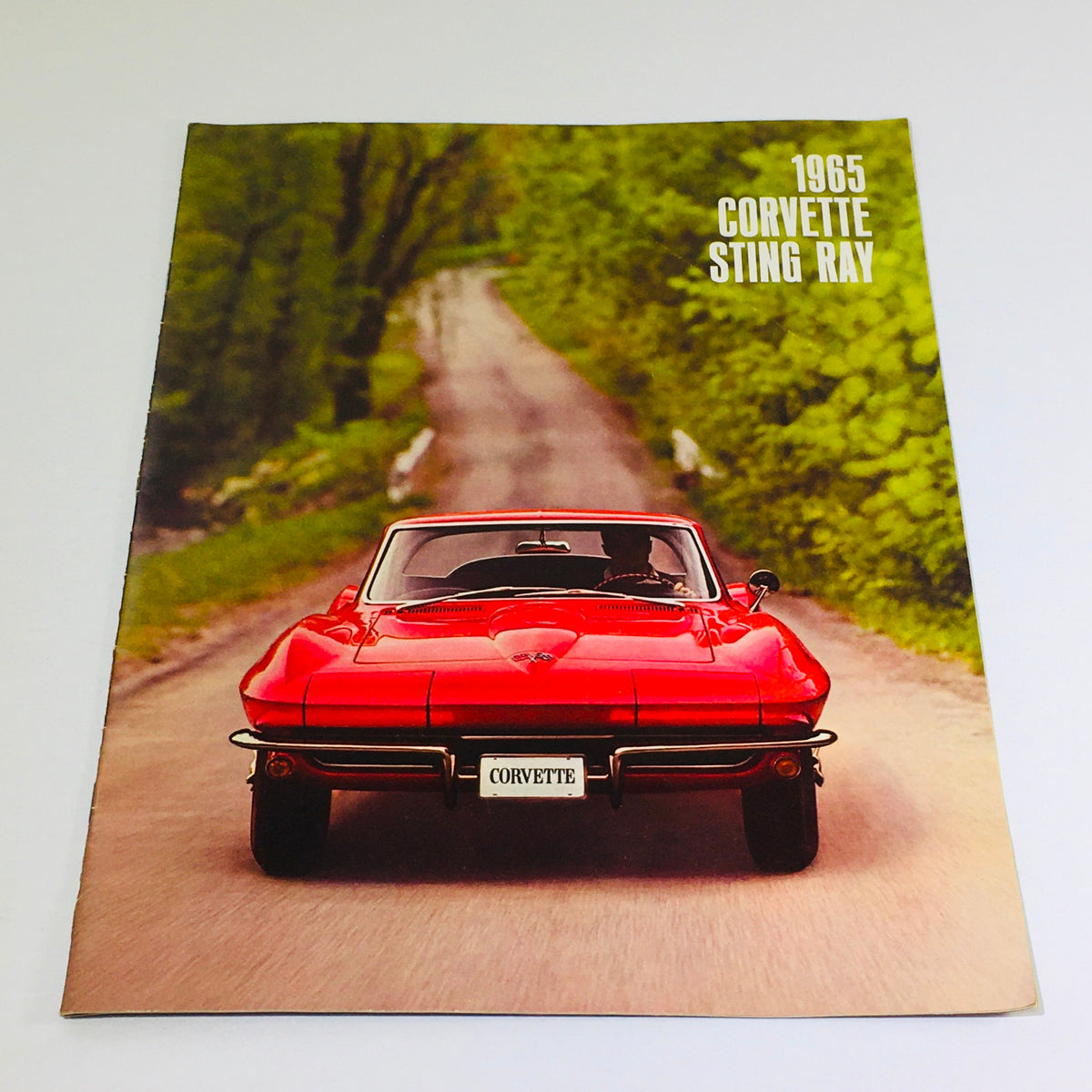 1965 Chevrolet Corvette Stingray Coupe brochure cover showcasing a red sports car on a scenic road, representing classic American automotive design.
