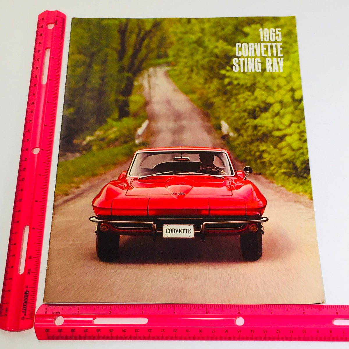 Close-up of the 1965 Chevrolet Corvette Stingray Coupe brochure cover with a ruler for scale, illustrating the iconic red sports car against a lush backdrop.