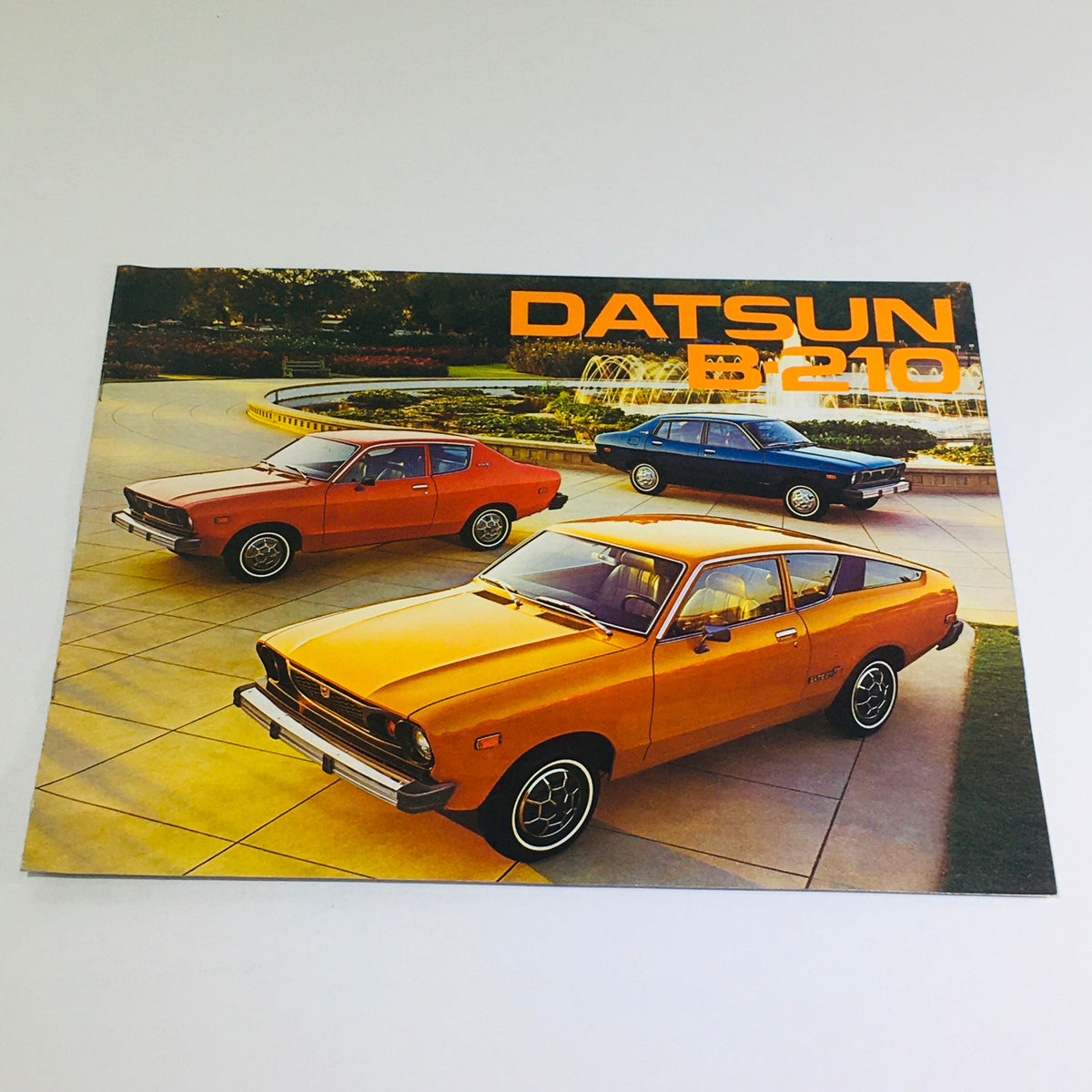 1975 Datsun B-210 Specifications Dealership Car Auto Brochure Catalog showcasing three cars, including an orange Datsun B-210, in a scenic outdoor setting.