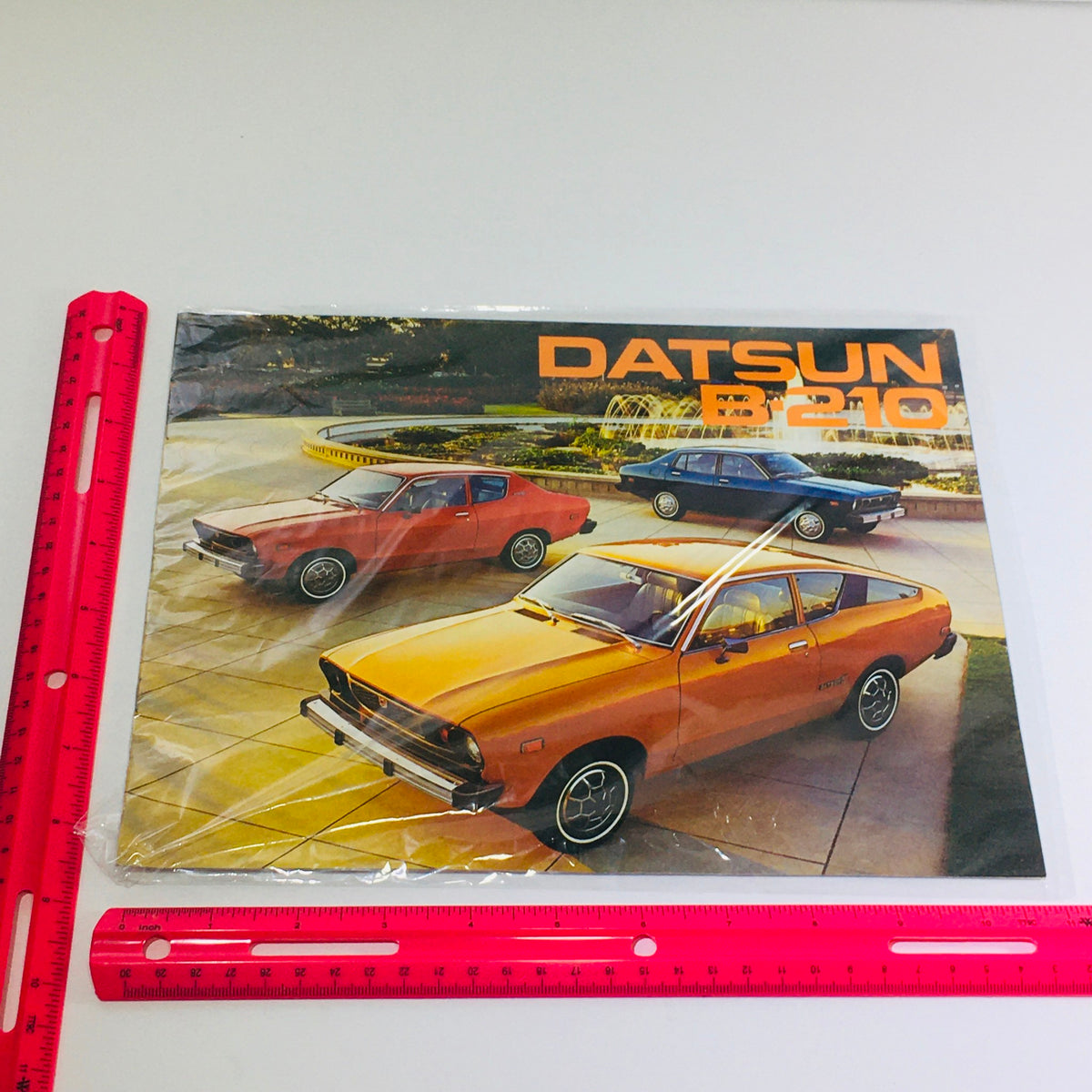 1975 Datsun B-210 Specifications Dealership Car Auto Brochure Catalog featuring an orange Datsun B-210 in the foreground, with two additional Datsun cars in the background. The scene is set in a well-maintained landscape, emphasizing the car's design and style.