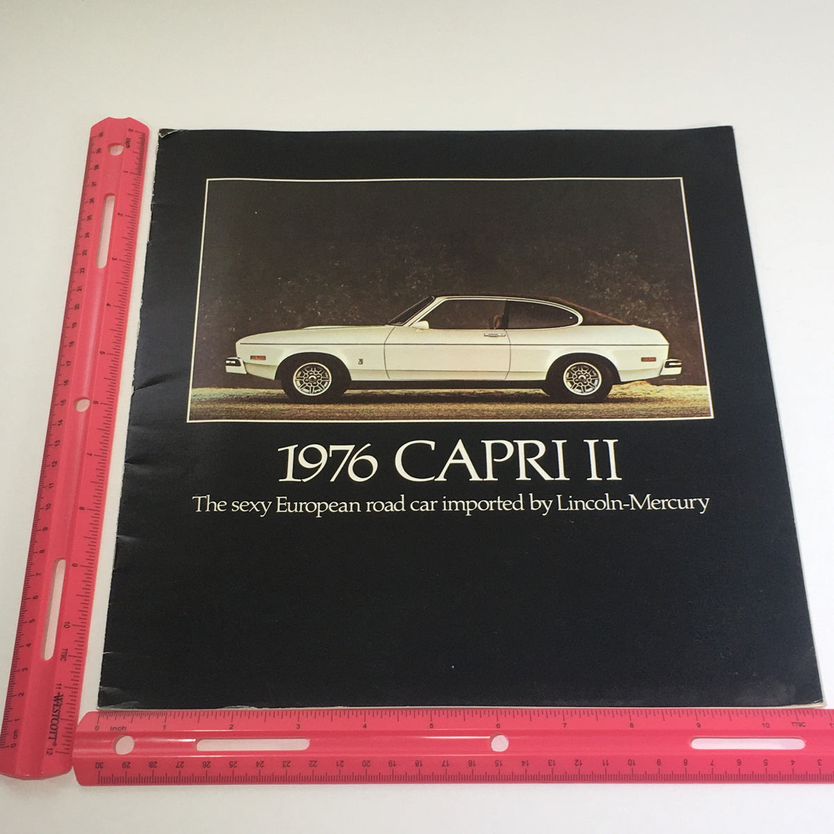 The front cover of the 1976 Lincoln-Mercury Division Capri II Dealership Car Auto Brochure showcasing a white Capri II model, accompanied by a ruler for size reference, emphasizing the classic European design of the vehicle.