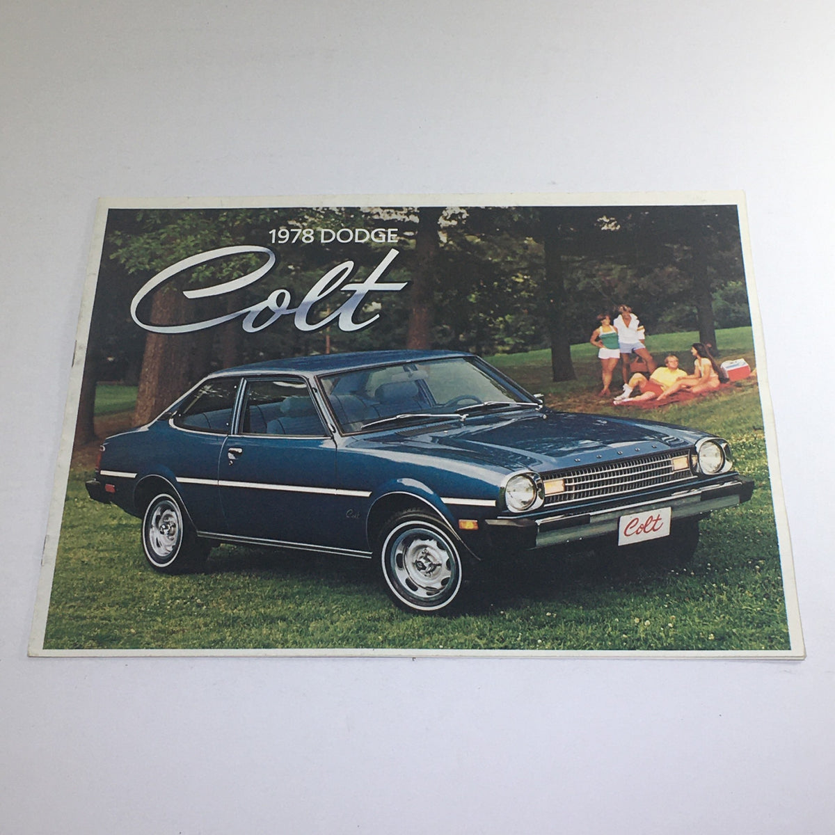 1978 Dodge Colt Custom Coupe brochure featuring the car parked on a grassy area with people enjoying a picnic in the background. This collectible catalog showcases the sleek lines and vintage design of the Dodge Colt, part of the 1978 Dodge Cold Custom Coupe M/M Dealership Car Auto Brochure.