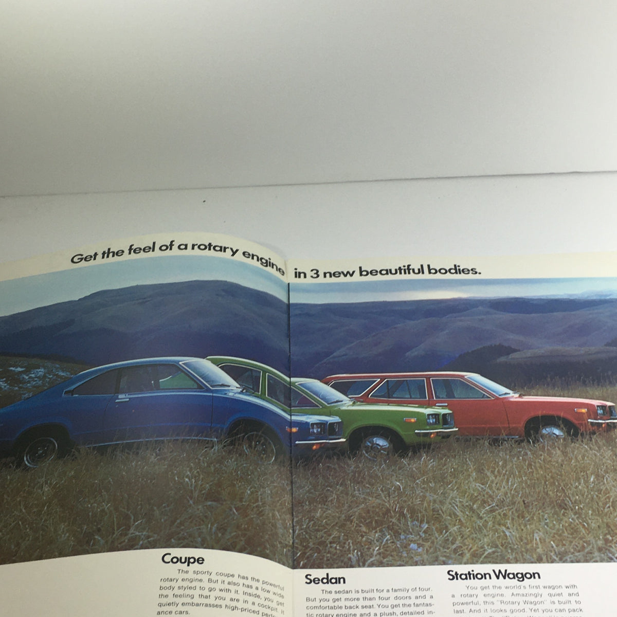 Inside layout of the 1973 Mazda RX-3 Coupe Sedan Station Wagon Dealership Car Auto Brochure showcasing a blue coupe, green and orange sedan, and a station wagon in a scenic field.