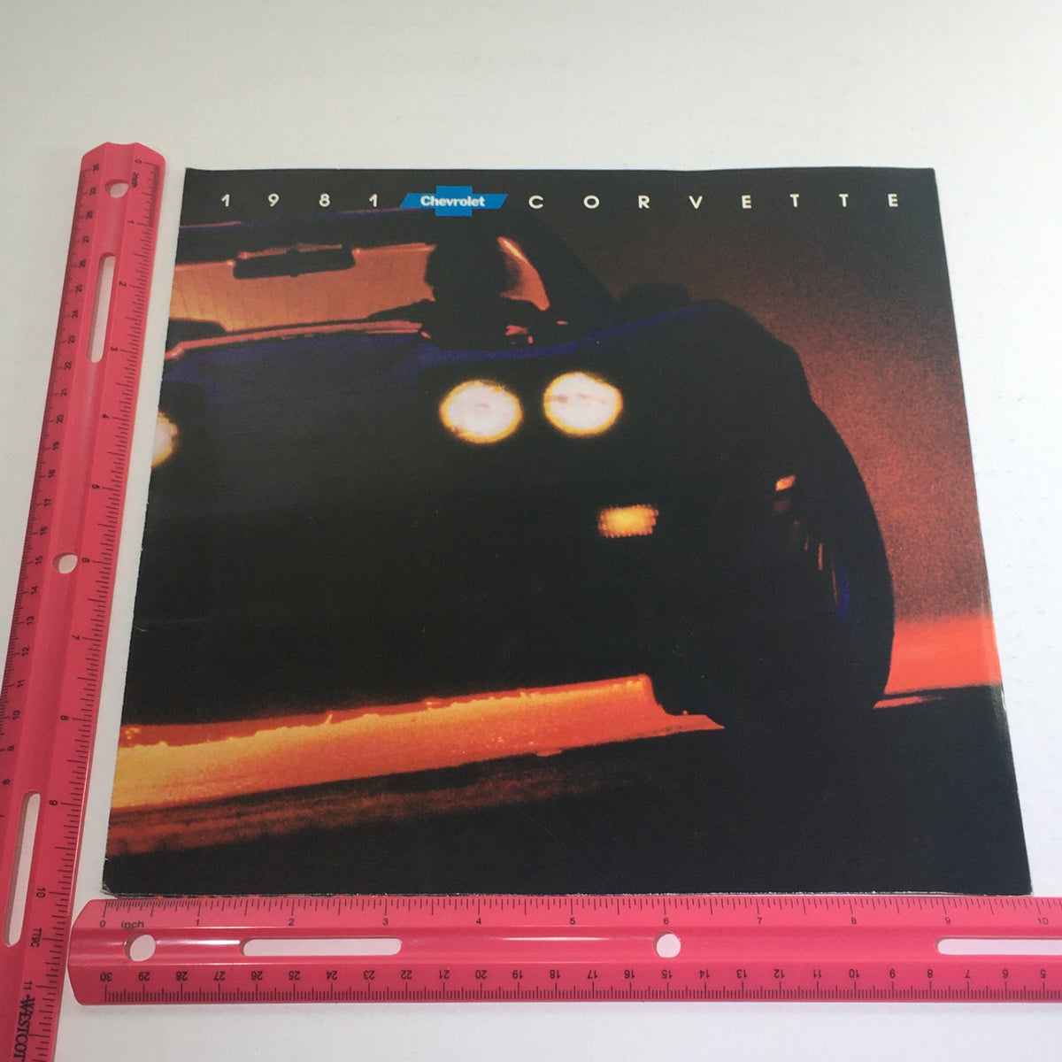 A 1981 Chevrolet Corvette Dealership Car Auto Brochure Catalog displayed next to a ruler for size reference, illustrating its dimensions.