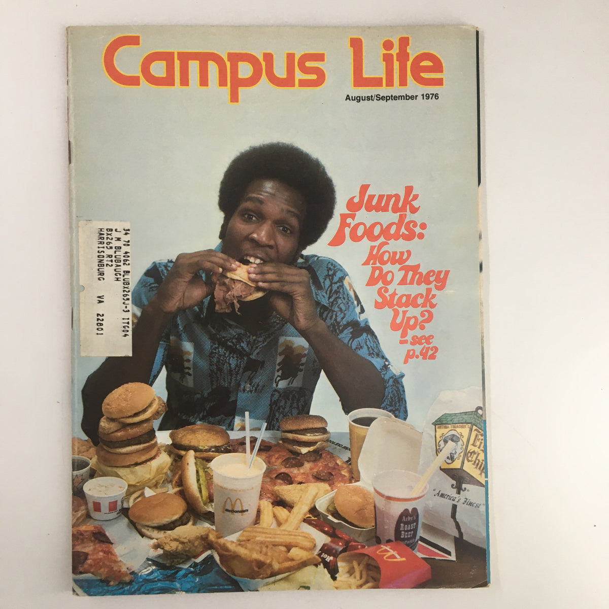 Cover of VTG Campus Life Magazine August 1976 featuring a person eating snacks surrounded by various fast food items, titled 'Junk Foods: How Do They Stack Up?'.