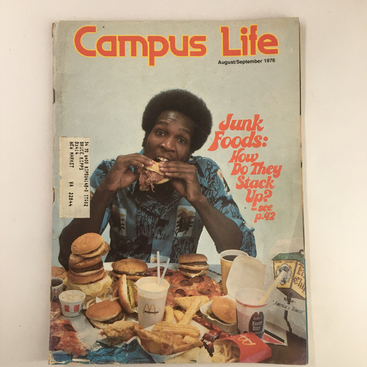 Cover of VTG Campus Life Magazine, August 1976, featuring an individual enjoying a burger surrounded by junk food, with the headline 'Junk Foods: How Do They Stack Up?'.