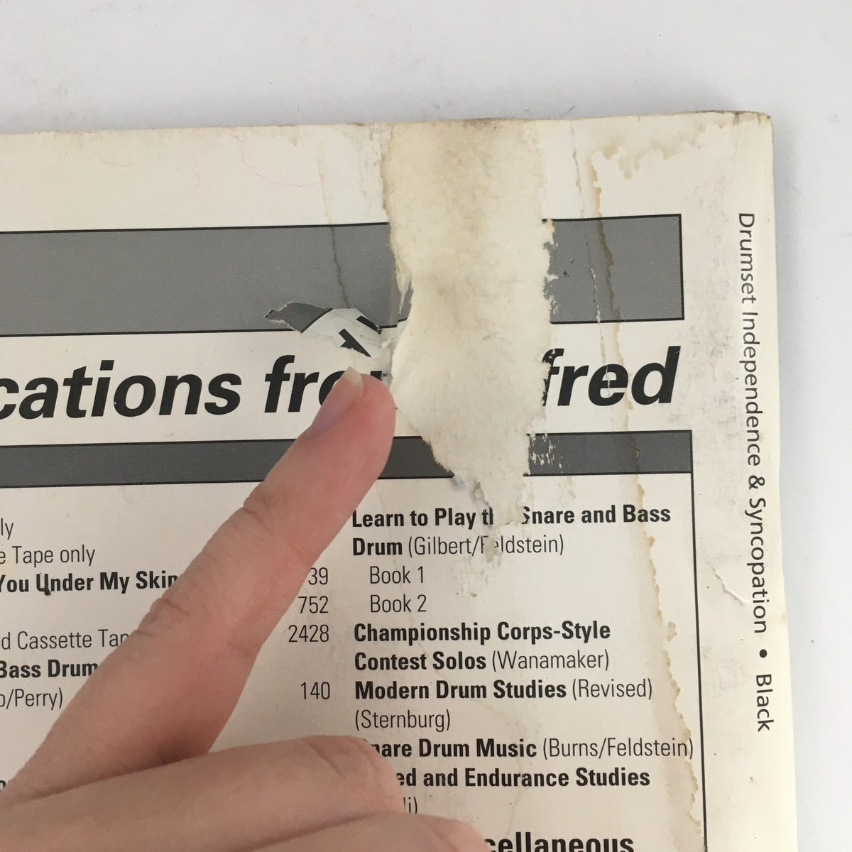 Close-up view of the back cover of 'Drumset Independence & Syncopation Rhythms by Dave Black & The Jazz Feels', showing minor wear and tear, with a finger pointing at a torn area.