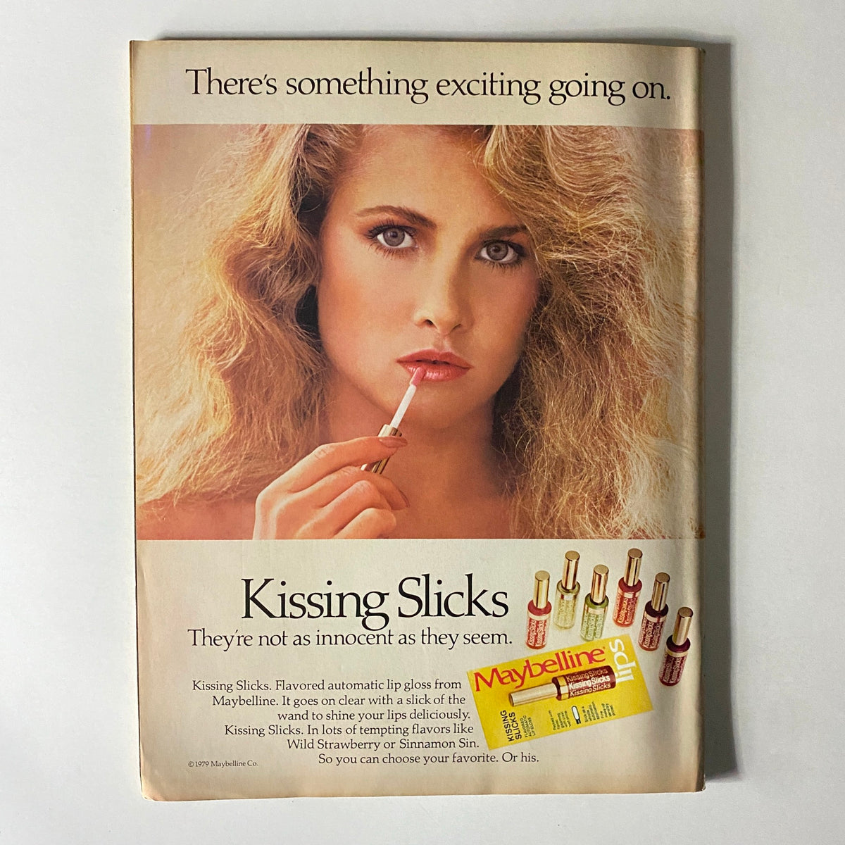 Vintage Maybelline advertisement for Kissing Slicks lip gloss, showcasing a model applying the gloss with a seductive pose, promoting 1980s beauty trends.