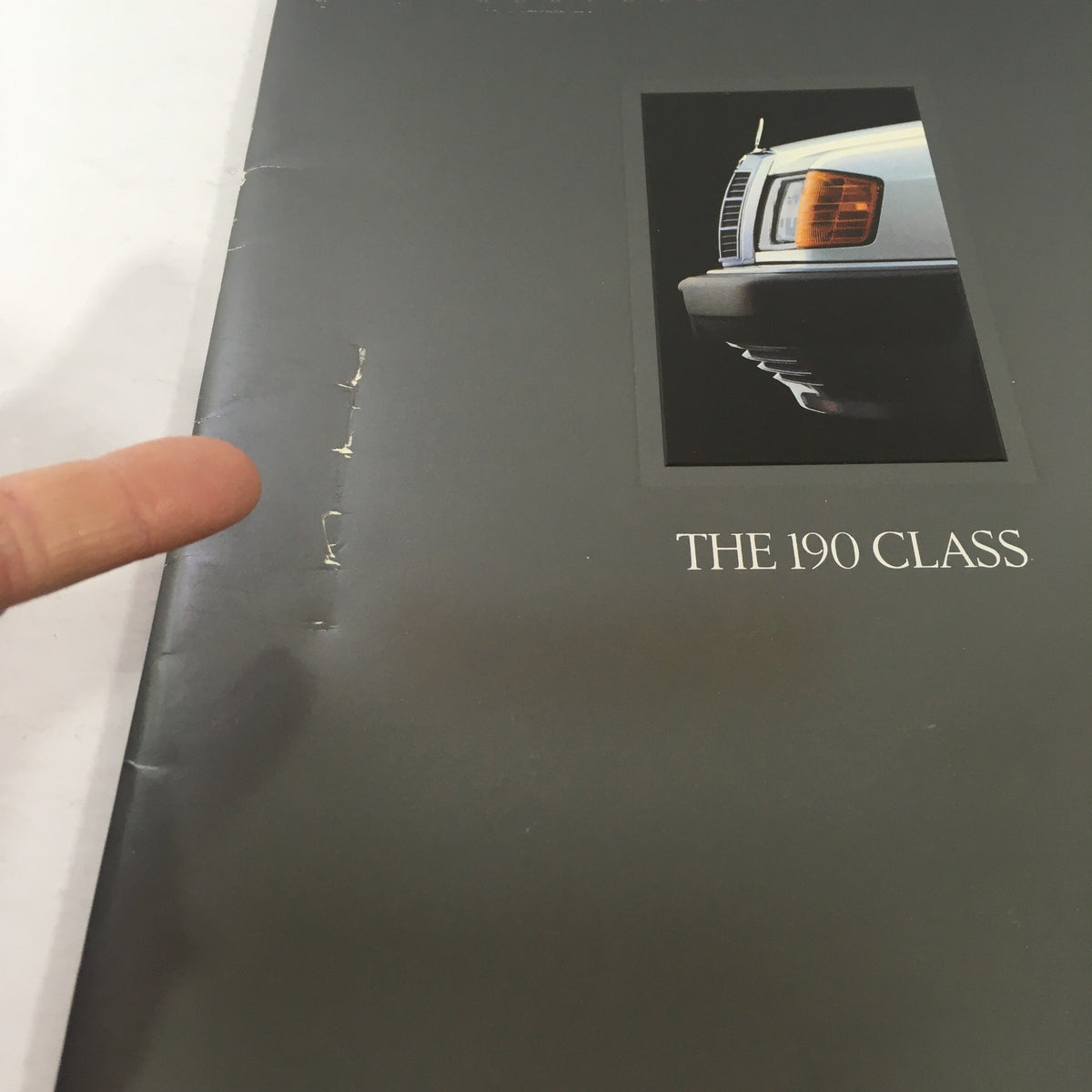 Detail of the 1989 Mercedes-Benz 190-Class 190E 2.6 Sedan Dealership Car Auto Brochure Catalog cover showing damage with a finger pointing at the tear, highlighting the catalog's used condition.