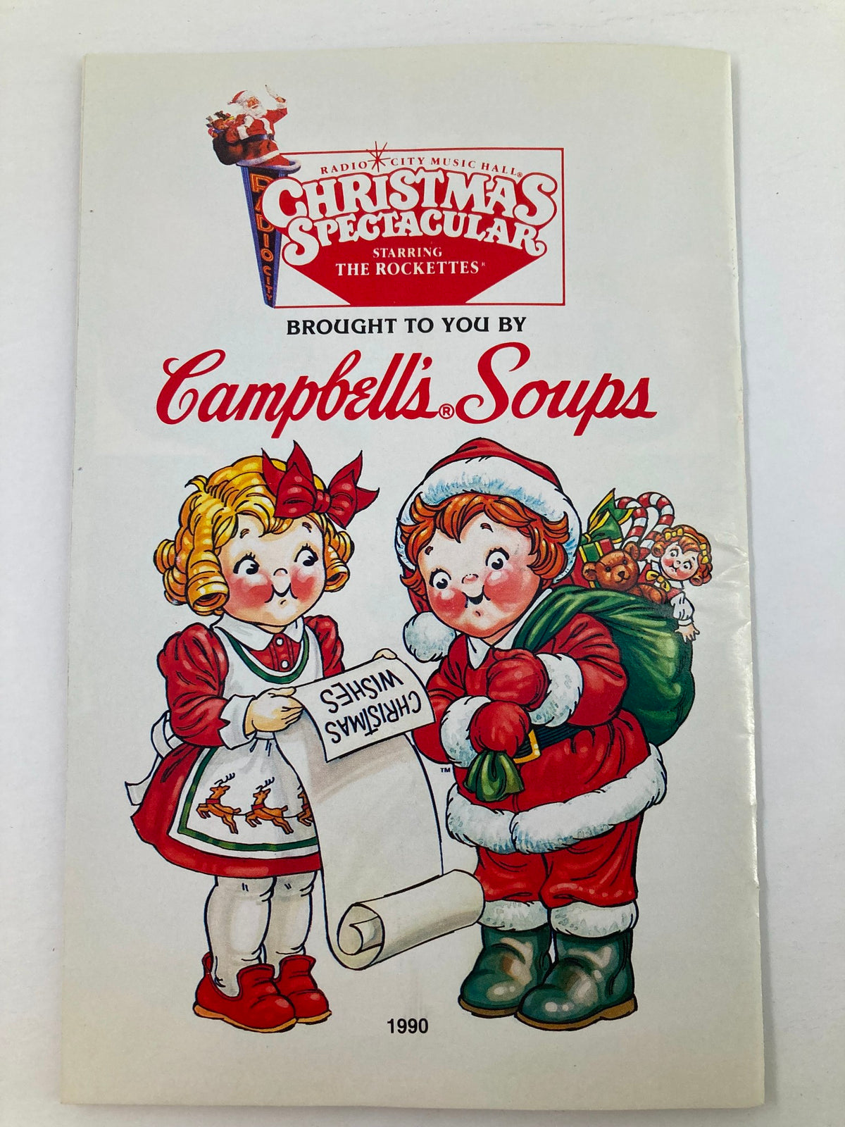 1990 Program Radio City Music Hall The Rockettes in Christmas Spectacular cover featuring festive illustrations of children with a Christmas wish list and a Santa bag, highlighting the joyful holiday spirit associated with the show.