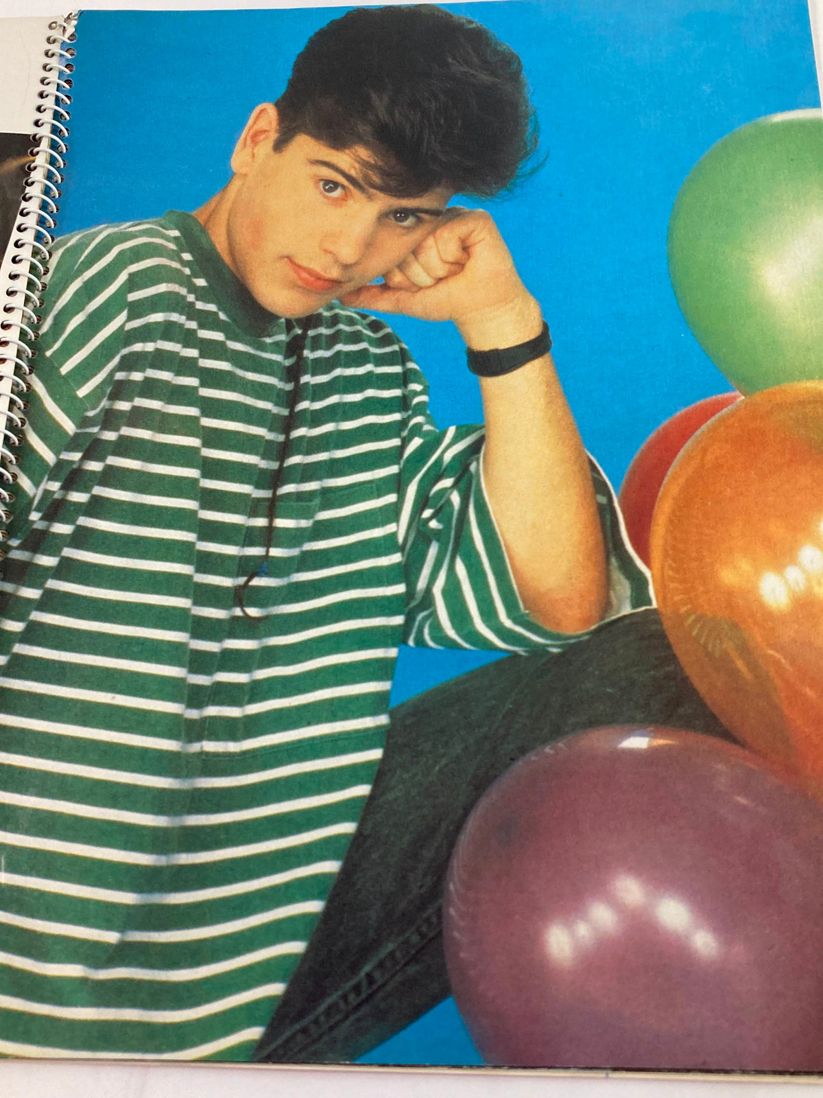 Image from '1990 Publications International, Ltd. Presents New Kids On The Block' showing Jonathan Knight in a casual striped outfit against a vibrant blue background, surrounded by colorful balloons.