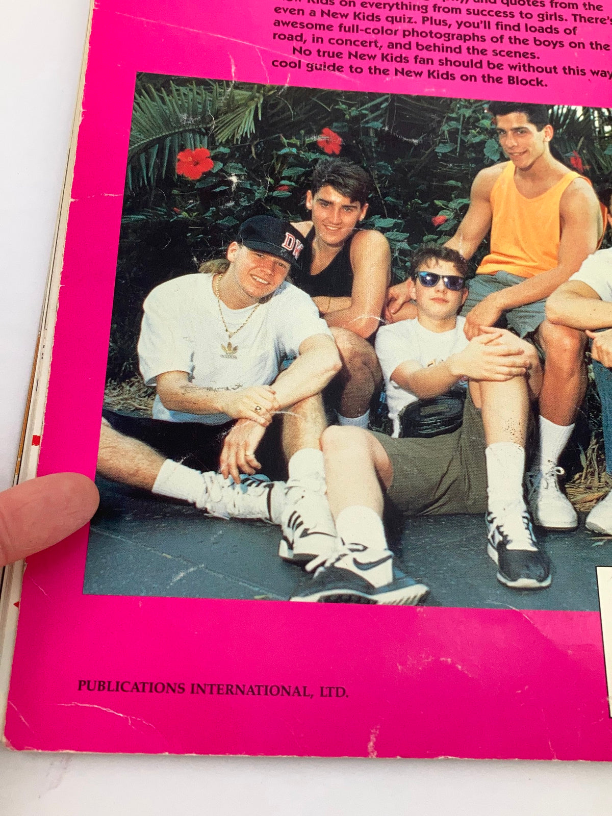 A group of young men sitting together, smiling, and enjoying a sunny day, showcasing the 1990 Publications International, Ltd. Presents New Kids On The Block magazine. The background includes lush greenery, complementing the vibrant pink cover of the publication.