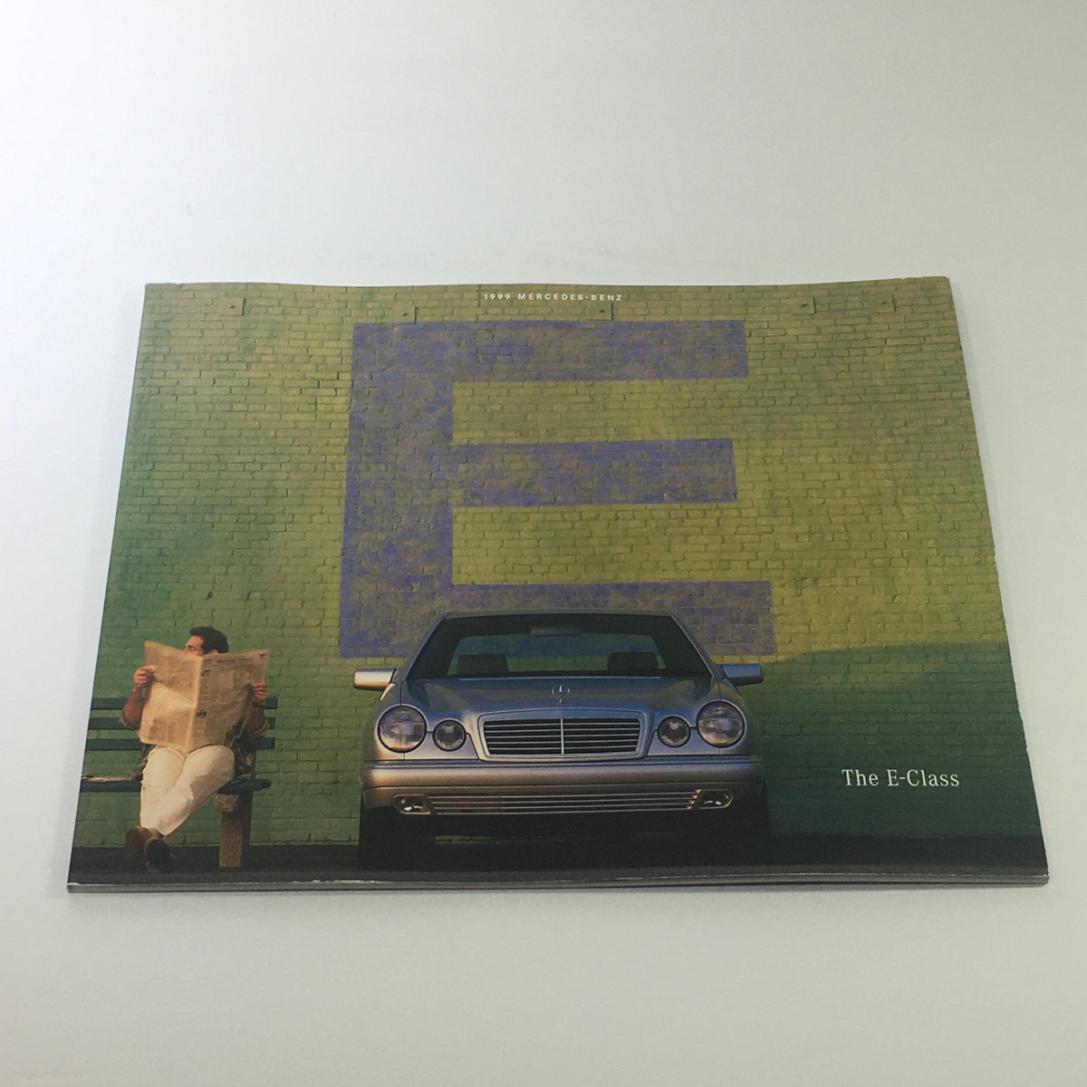 1999 Mercedes-Benz E-Class Dealership Car Auto Brochure Catalog with a man reading a newspaper in front of a silver Mercedes-Benz E-Class against a colorful brick wall.