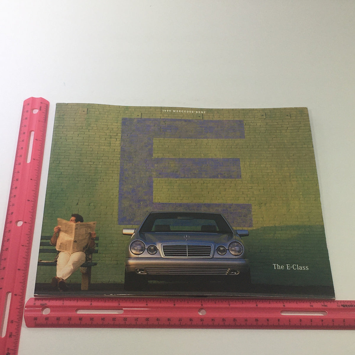 Front cover of the 1999 Mercedes-Benz E-Class Dealership Car Auto Brochure Catalog featuring a silver E-Class parked alongside a person reading a newspaper.