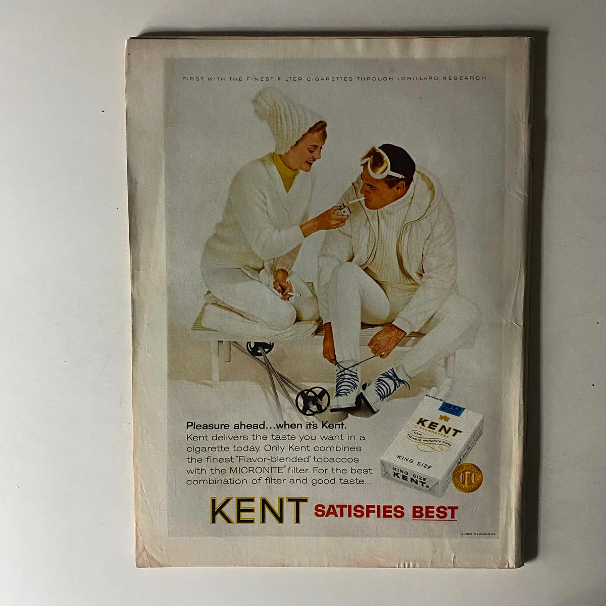 Vintage advertisement for Kent cigarettes from a 1965 magazine, featuring a couple engaging in winter sports, promoting the quality of Kent cigarettes with a focus on the MICRONITE filter.