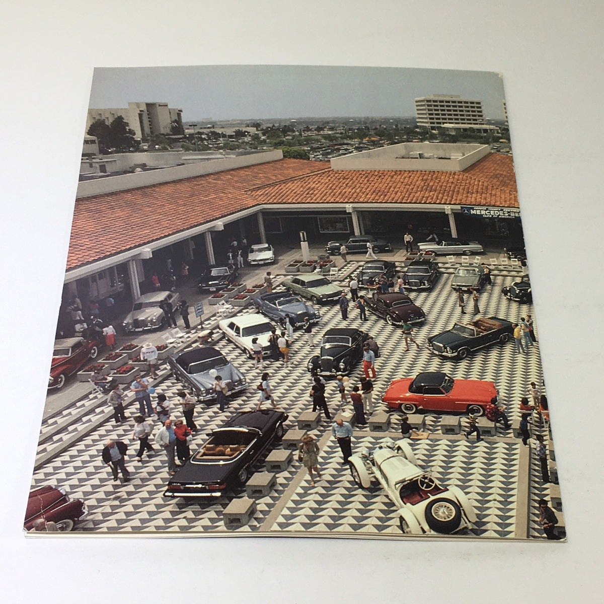 Exterior view of a Mercedes-Benz dealership from the 1980s, as seen in the 1982 Mercedes-Benz Volume VI Dealership Car Auto Brochure Catalog, showcasing classic cars and people around.