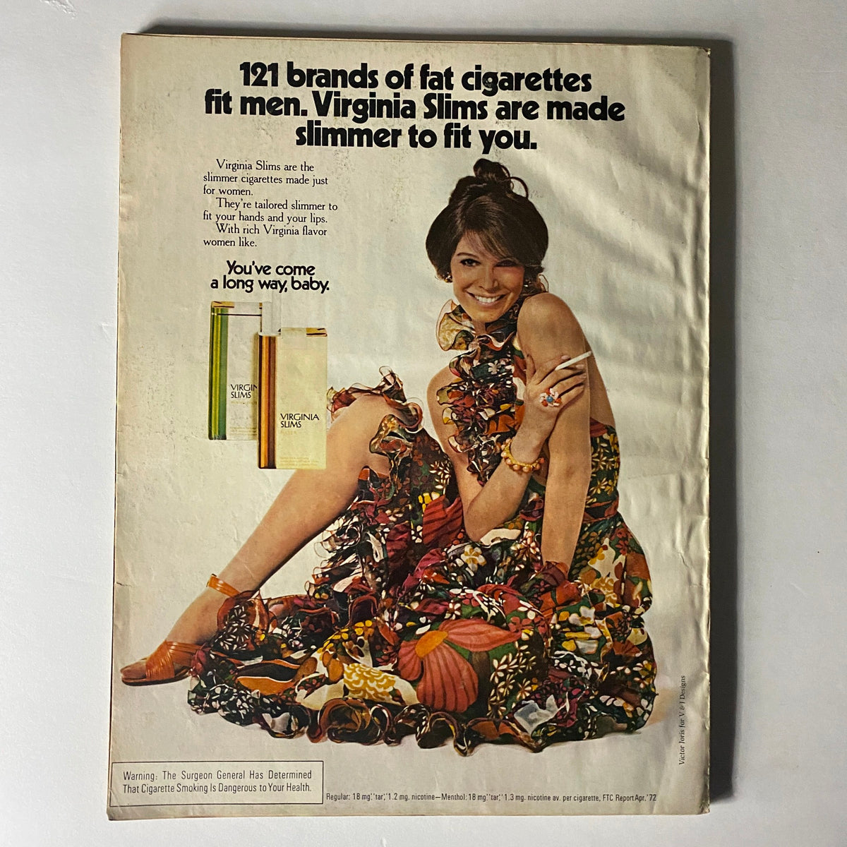 Advertisement featuring Virginia Slims cigarettes with the tagline 'You've come a long way, baby'. The ad showcases a stylish woman in a colorful floral wrap dress, promoting the brand as a slimmer cigarette option tailored for women. This image relates to the iconic Harper's Bazaar Magazine October 1972 Ali MacGraw Cover.