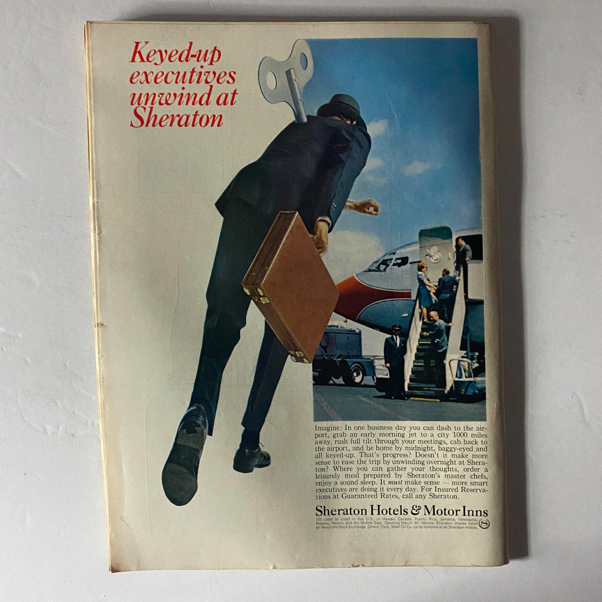 Advertisement from a vintage magazine promoting Sheraton Hotels, featuring a businessman with a briefcase and a whimsical wind-up key, emphasizing relaxation for busy executives.