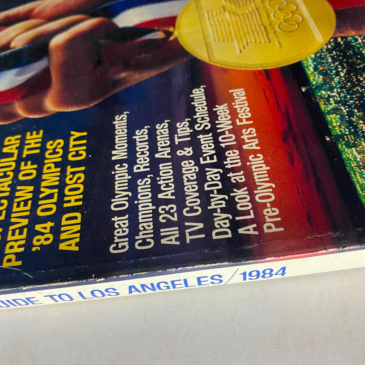 Close-up view of the cover of the 1984 Official Olympic Guide To Los Angeles Collector's Edition, focusing on promotional text highlighting Olympic moments and event schedules.