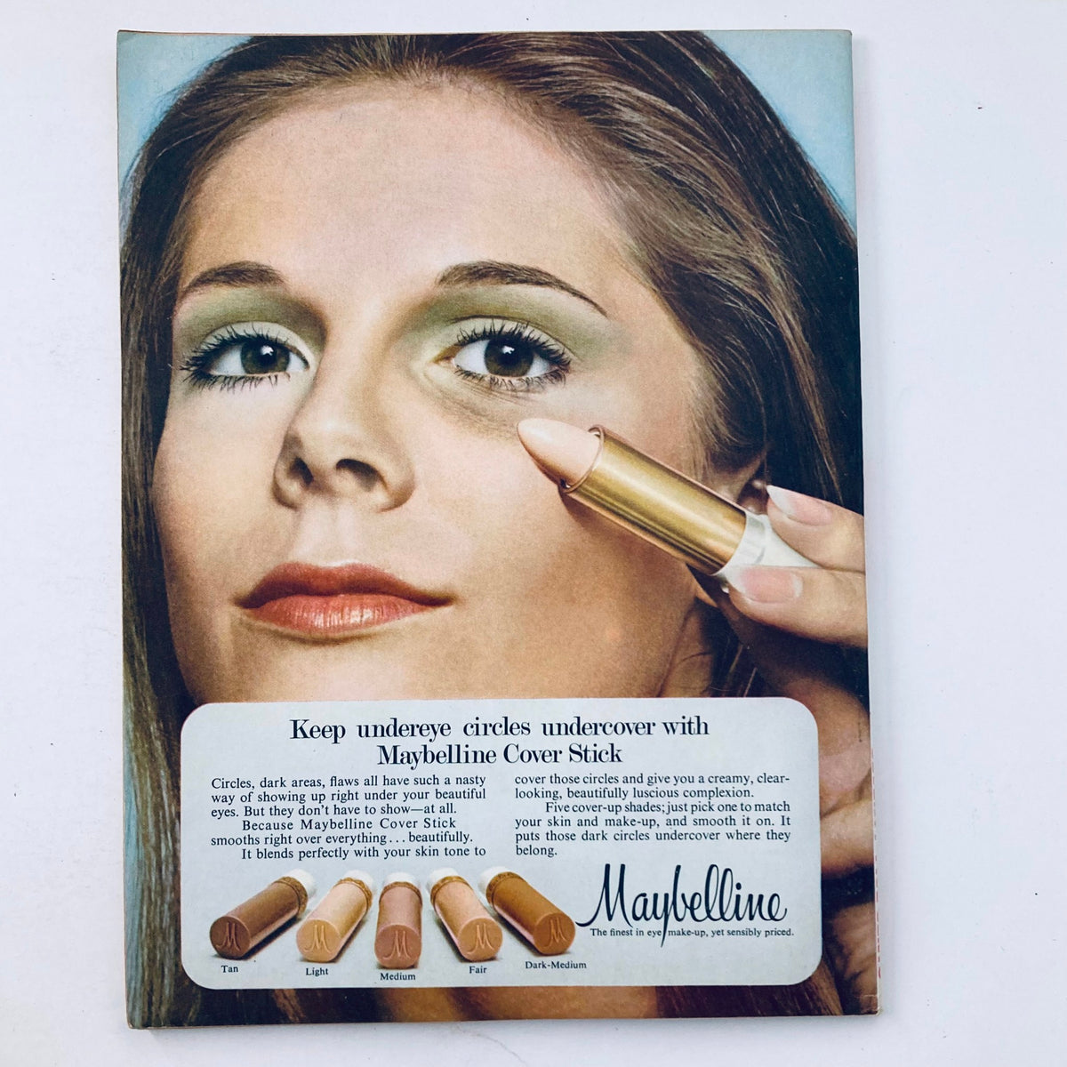 Vintage Maybelline advertisement featuring a model demonstrating the Maybelline Cover Stick used for concealing under-eye circles, accompanied by color options. Ideal for collectors of retro beauty products.