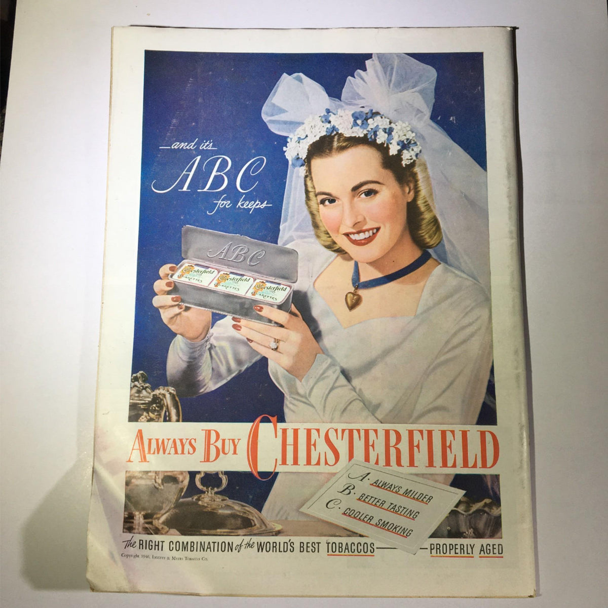 Advertisement for Chesterfield cigarettes from a vintage magazine, featuring a smiling woman showcasing cigarette packs, set against a blue background.
