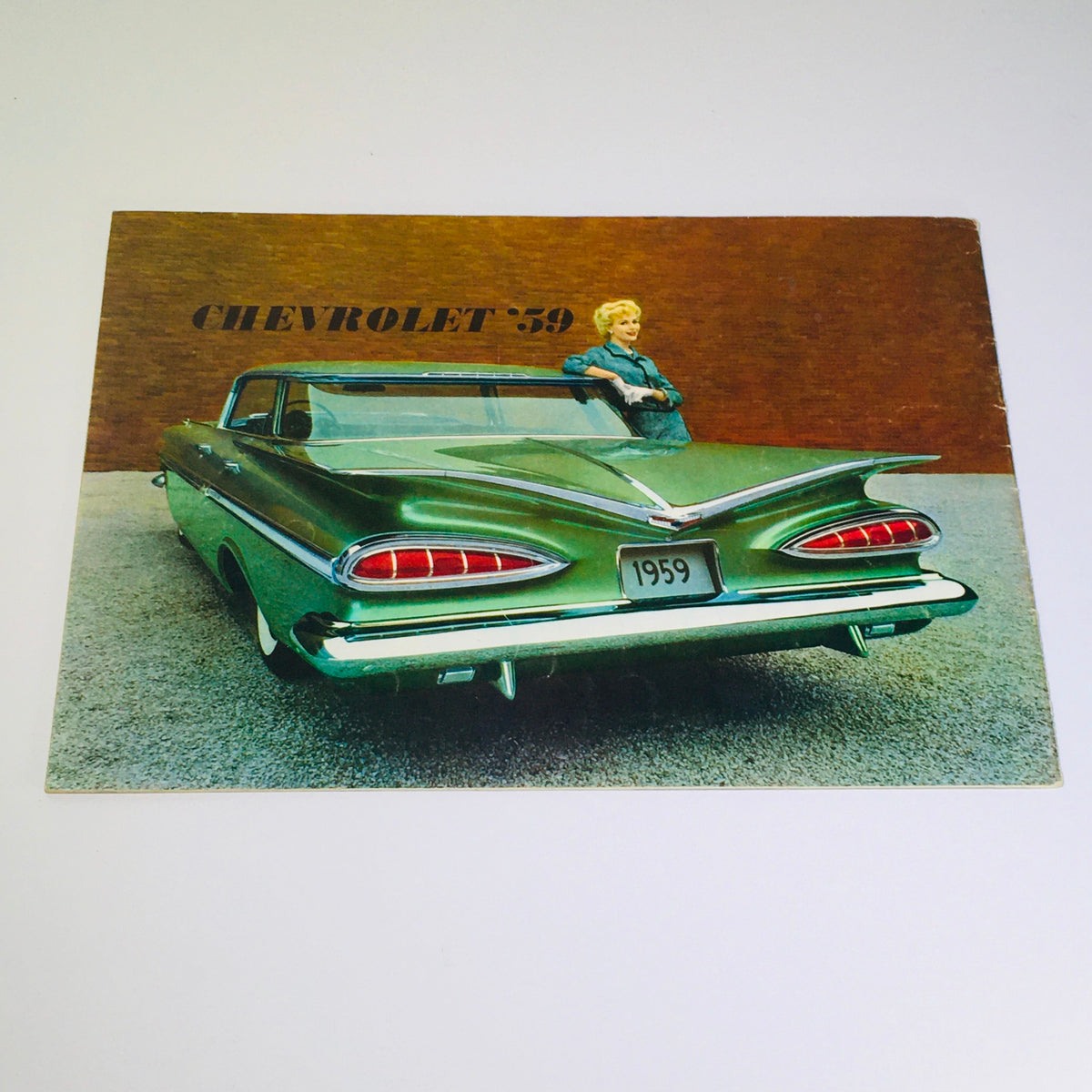 Back view of the Vintage 1959 Chevrolet Kingswood 4-Door Station Wagon Hardtop Car Catalog Brochure showcasing a green car with prominent tail lights and a woman leaning on it.