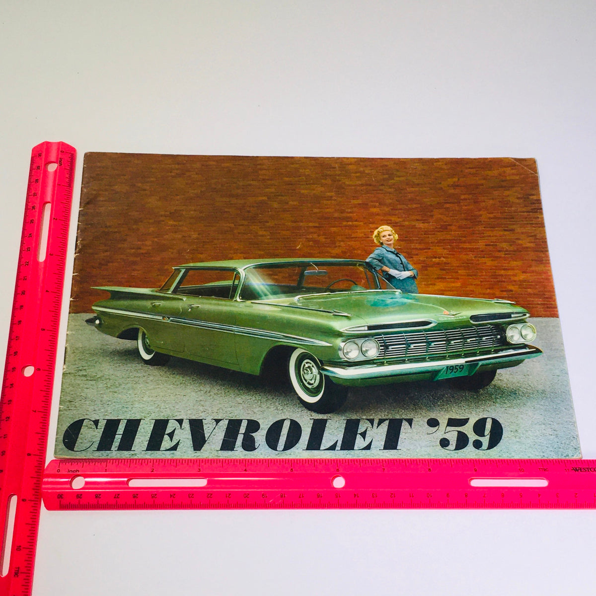Vintage 1959 Chevrolet Kingswood 4-Door Station Wagon Hardtop Car Catalog Brochure featuring a classic green Chevrolet car with a woman in vintage attire, set against a brick wall backdrop, showcasing its design and style.