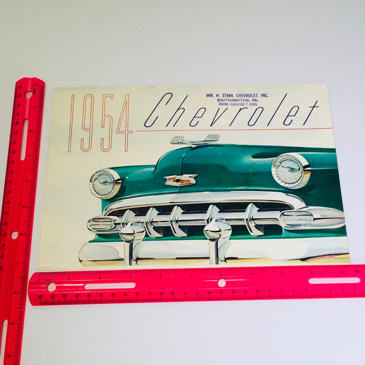 Close-up of the cover of the Vintage 1954 Chevrolet Bel-Air 4-Door Sedan Blue-Flame 125 Engine Car Brochure with a ruler for scale, displaying the classic styling and color of the brochure.