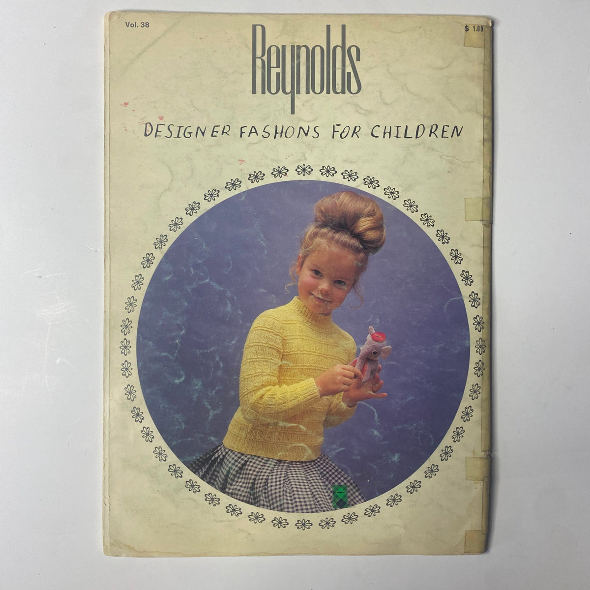 Cover of the 1980s Reynolds Designer Fashions for Children Catalog featuring a girl in a yellow sweater holding a pink toy, showcasing vintage children's fashion.