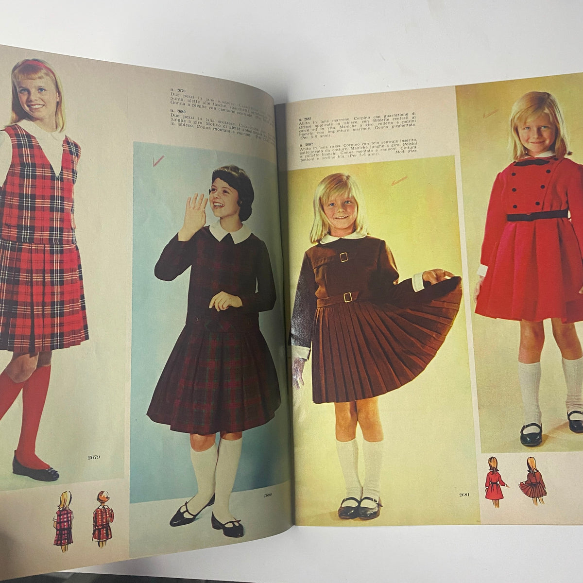 1962 La Moda Dei Bimbi Autumn and Winter Catalog featuring four stylish girls' dresses in vibrant colors and patterns. The left page displays two plaid dresses, while the right showcases a red dress and a classic brown outfit with a pleated skirt.