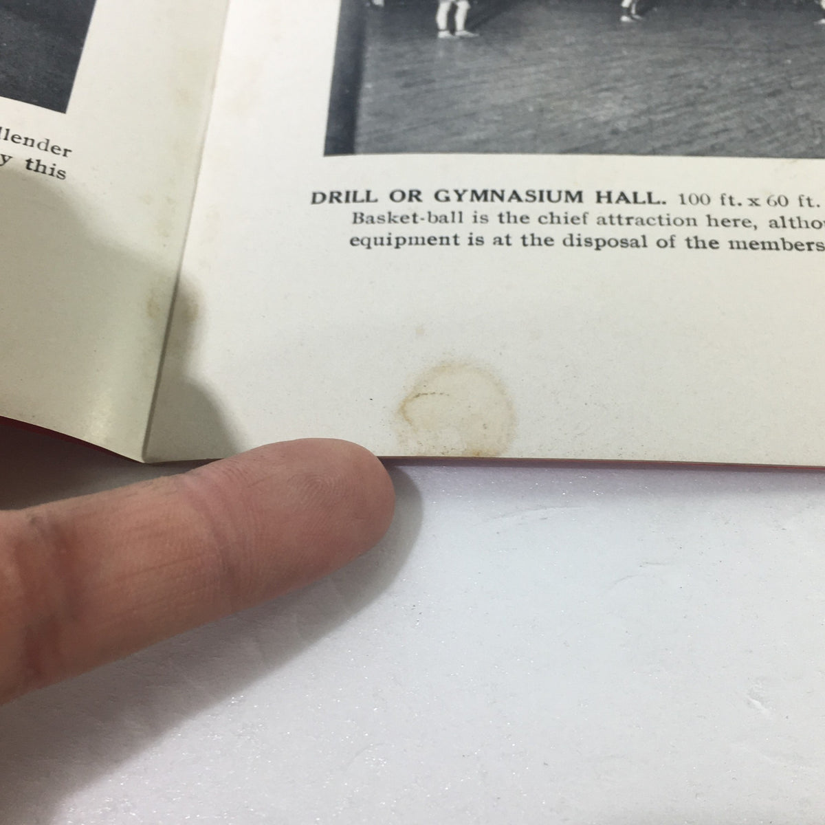 Close-up of a page from the VTG January 21, 1910 Military Education and Sports Armory of Company 'M' document, showing a description of the drill or gymnasium hall with a finger pointing to a minor stain on the page.
