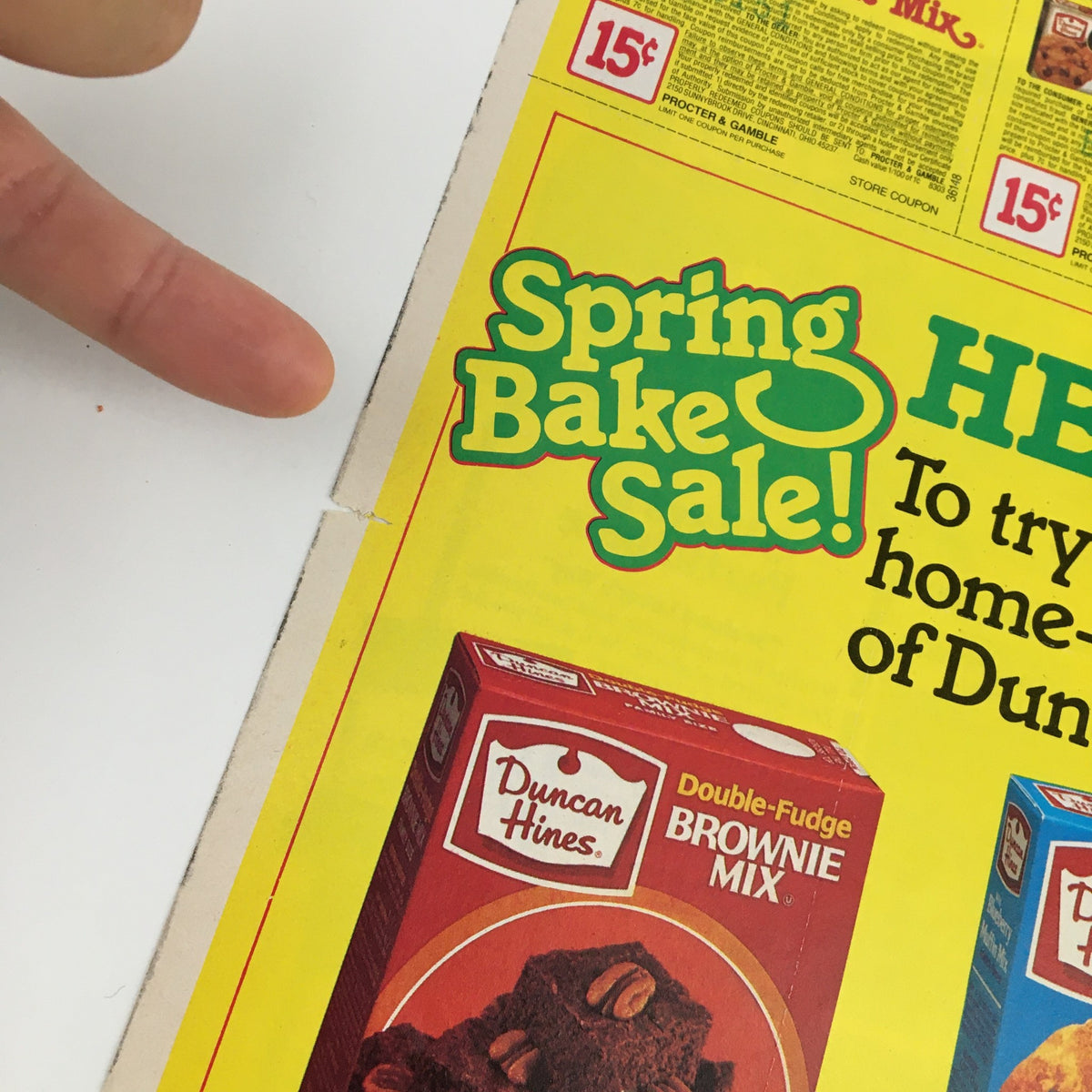 Close-up of the 1980 Duncan Hines Spring Bake Sale Circular Coupon Advertisement highlighting the 'Spring Bake Sale!' title and the Double-Fudge Brownie Mix, with finger pointing at the advertisement.