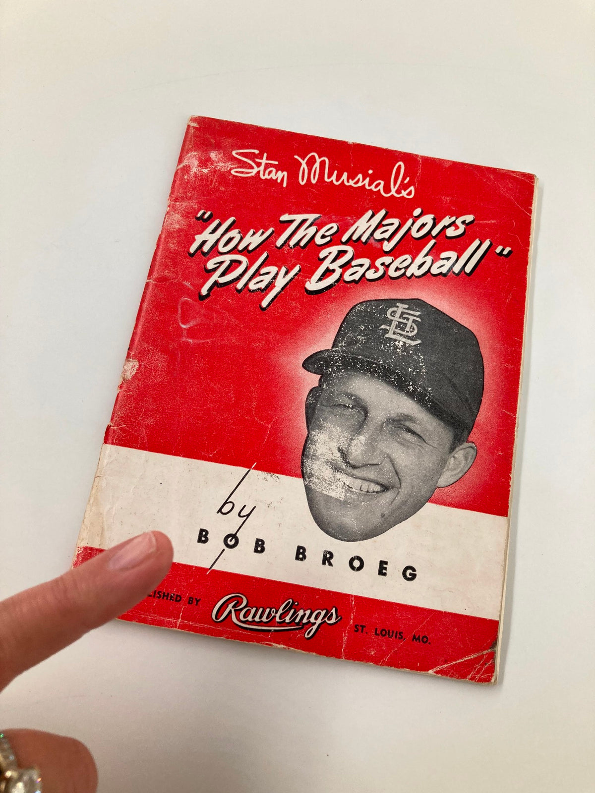 Close-up of the cover of 'Stan Musial's How The Majors Play Baseball' by Bob Broeg with a finger pointing at it, showcasing the book's vintage appeal.