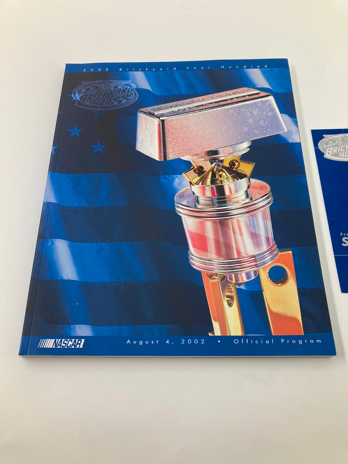 A close-up view of the NASCAR Brickyard 400 Official Program cover dated August 4 2002 with a vibrant trophy and flag design.