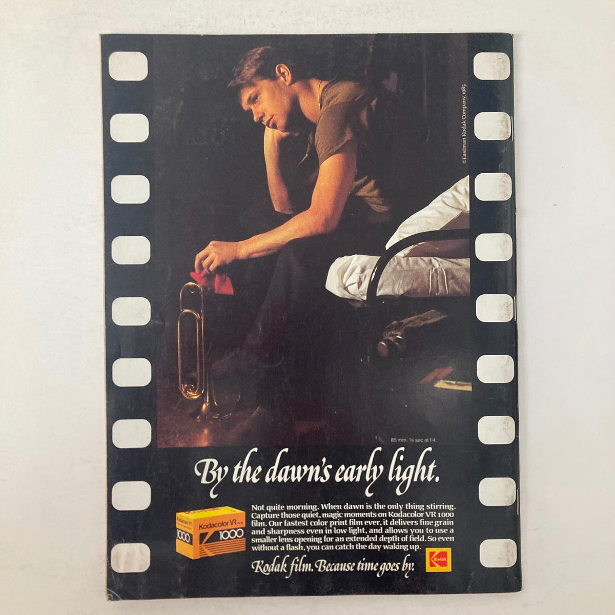 An advertisement from American Photographer Magazine January 1986, featuring a thoughtful young man with a trumpet in a dimly lit room, conveying the message 'By the dawn’s early light.' The ad promotes Kodak film, emphasizing its ability to capture moments in low light.