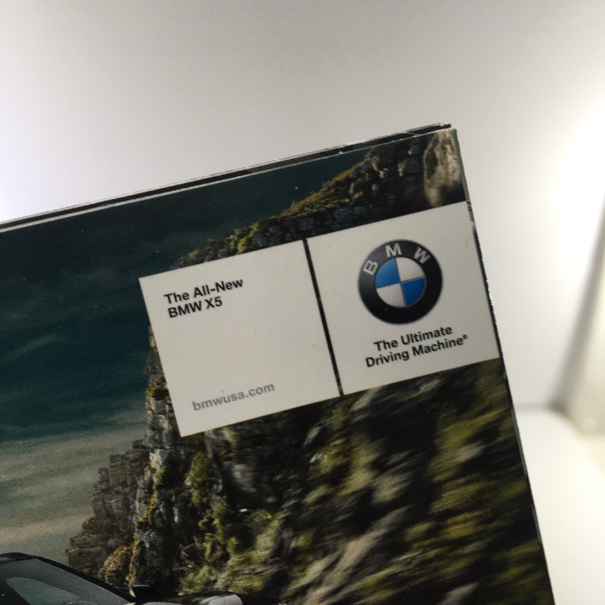 Close-up of the corner of the 2014 BMW X-5 Dealership Car Auto Brochure Catalog featuring the BMW logo and tagline 'The Ultimate Driving Machine'.