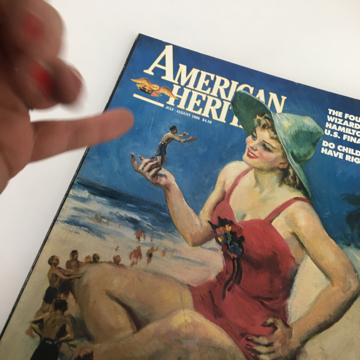 Close-up of the cover of American Heritage Magazine July 1990, showcasing a woman in a red swimsuit playfully holding a tiny figure, with beachgoers in the background and titles regarding Hamilton and U.S. Finance.